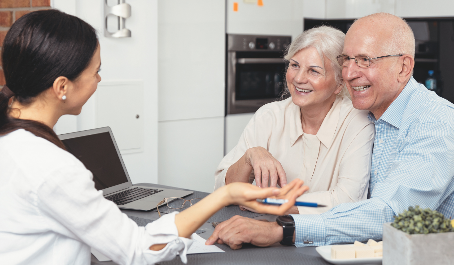 elderly husband and wife sitting side by side holding each other close and smiling to a director talking to them about senior care options